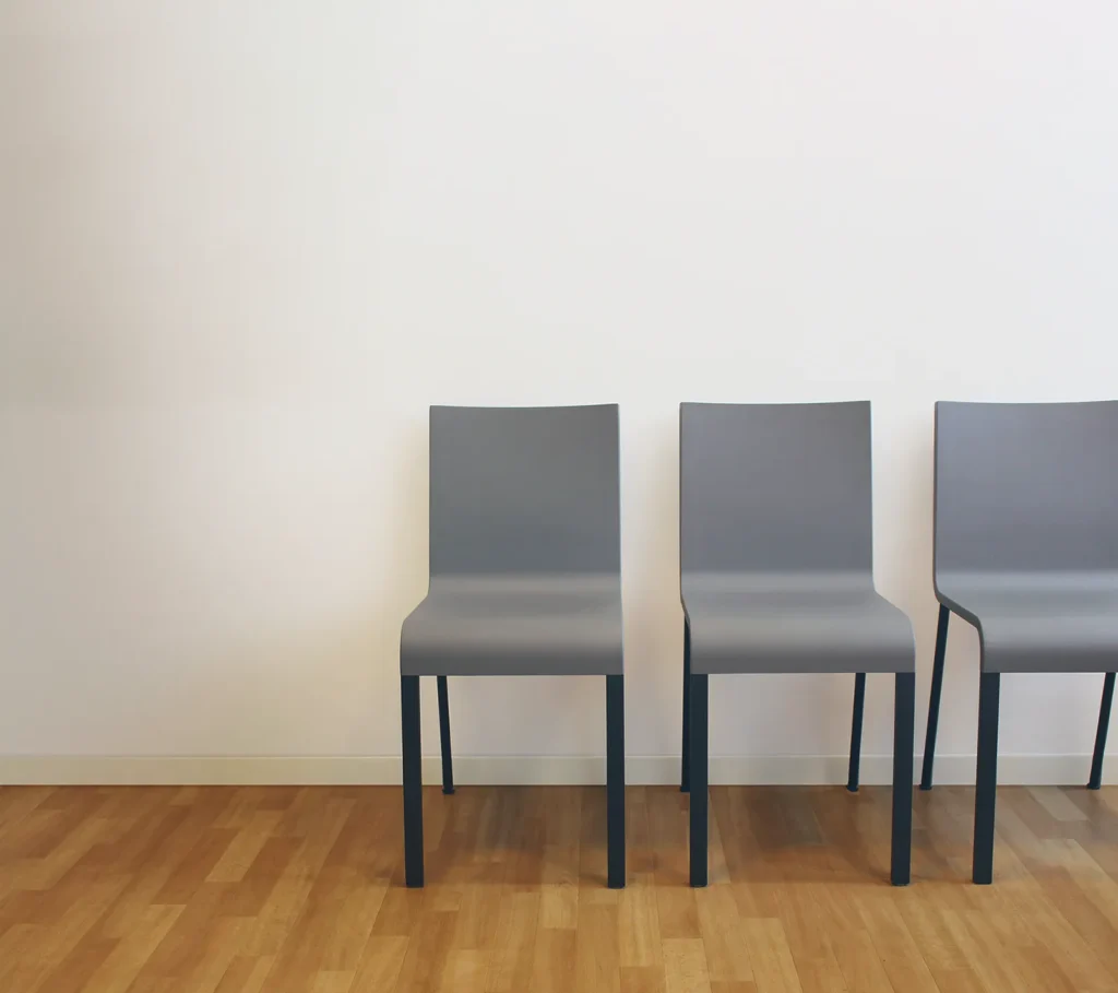 Drei graue Stühle aus dem Wartezimmer stehen nebeneinander aufgereiht vor einer weißen Wand. Der braune Parkettboden ist zu sehen.