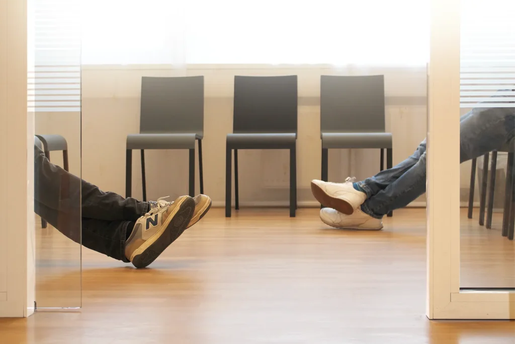 Das Wartezimmer der Praxis ist hell und die grauen Stühle wirken modern und aufgeräumt. Hier sitzen zwei Patienten, von denen man die Beine nach vorn ausgestreckt sieht.