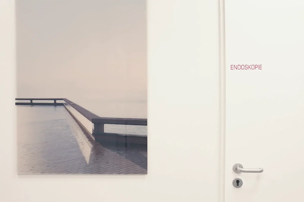Ein Bild aus der PRaxis das den Praxisflur zeigt von dem aus es zum Untersuchungsraum geht in der Endoskopien durchgeführt werden. Die Tür wird durch einen roten Schriftzug "Endoskopie" gekennzeichnet und daneben ist ein ruhiges Bild einer Küstenszene mit Blick aufs Meer.